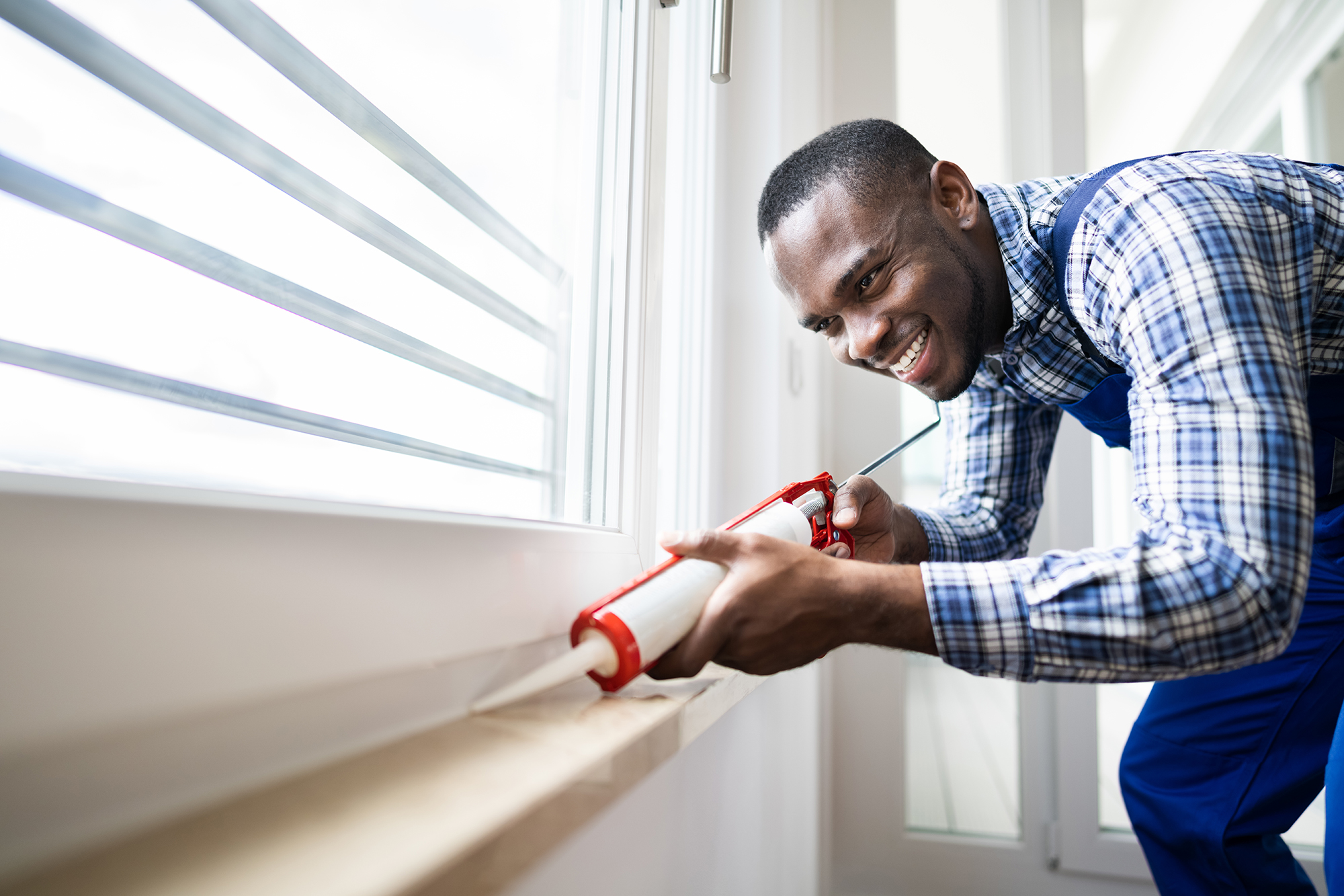 Winterizing doors and windows can reduce energy costs and keep you warmer. In this image, a man is caulking a window frame.